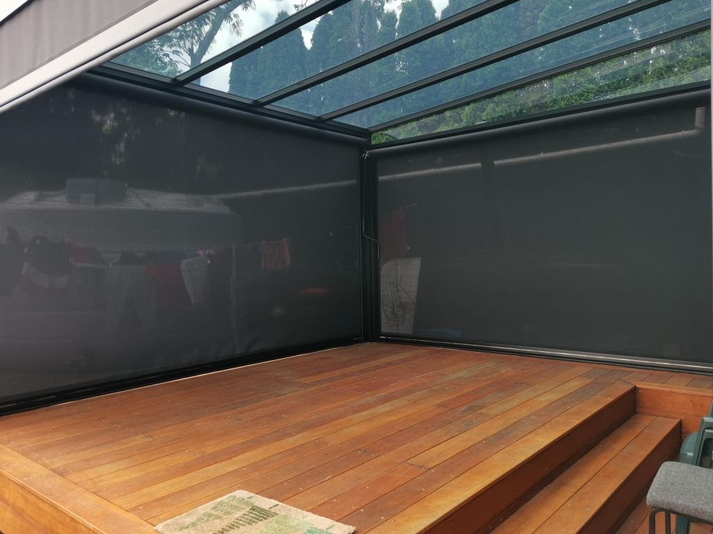 Interior view of a glass-roofed outdoor room with screen blinds and timber deck